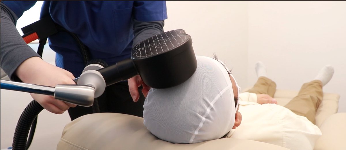 rTMSの施術の様子。リクライニングチェアで横になった方の頭部に、スタッフが磁気コイルを当てている。クリックするとYouTubeで動画を見られます。